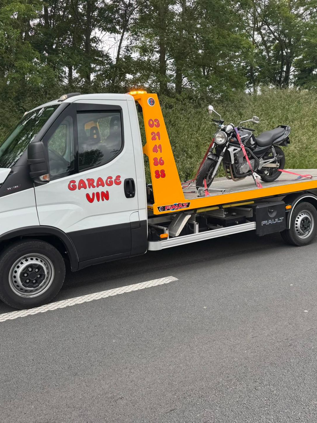 Moto sur une dépanneuse. Camion blanc, moto noire, en bord de route. Inscription sur le camion : GARAGE VIN avec un numéro de téléphone.