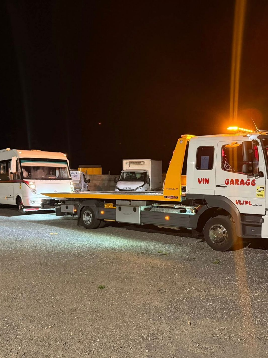 Une dépanneuse remorque une caravane blanche de nuit. Un autre camion est visible à l'arrière-plan. Les gyrophares jaunes sont allumés.