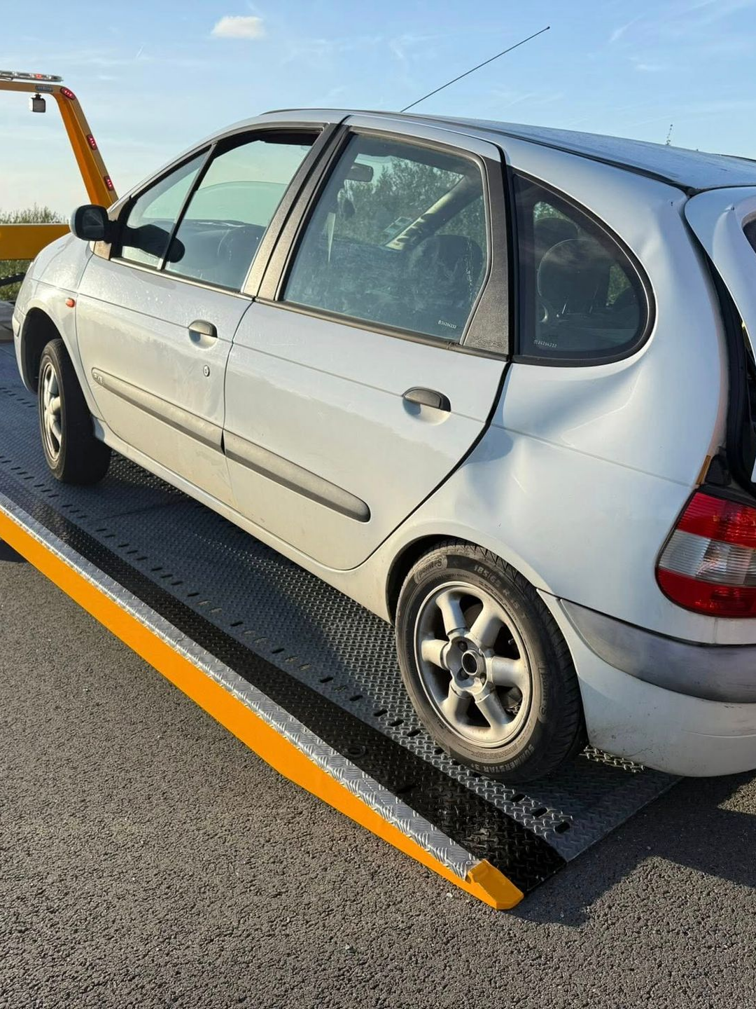 Voiture argentée sur une dépanneuse, aile arrière endommagée, stationnée sur l'asphalte.