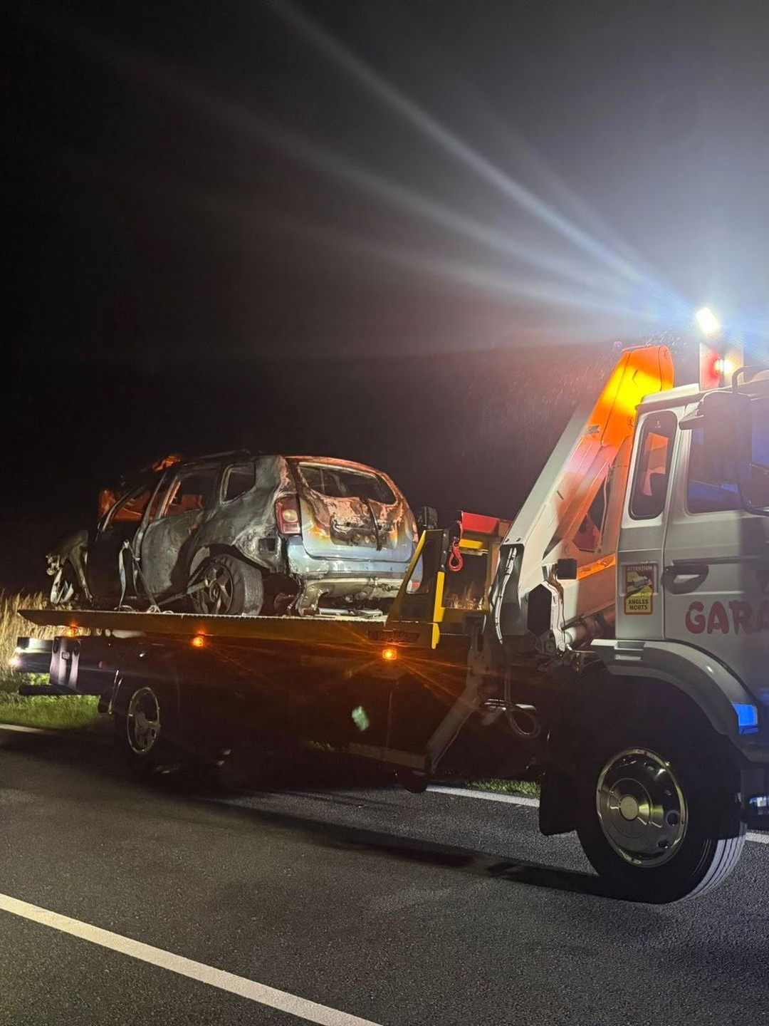 Une voiture calcinée chargée sur une dépanneuse, de nuit. Des gyrophares orange et bleu éclairent la scène sur une route.