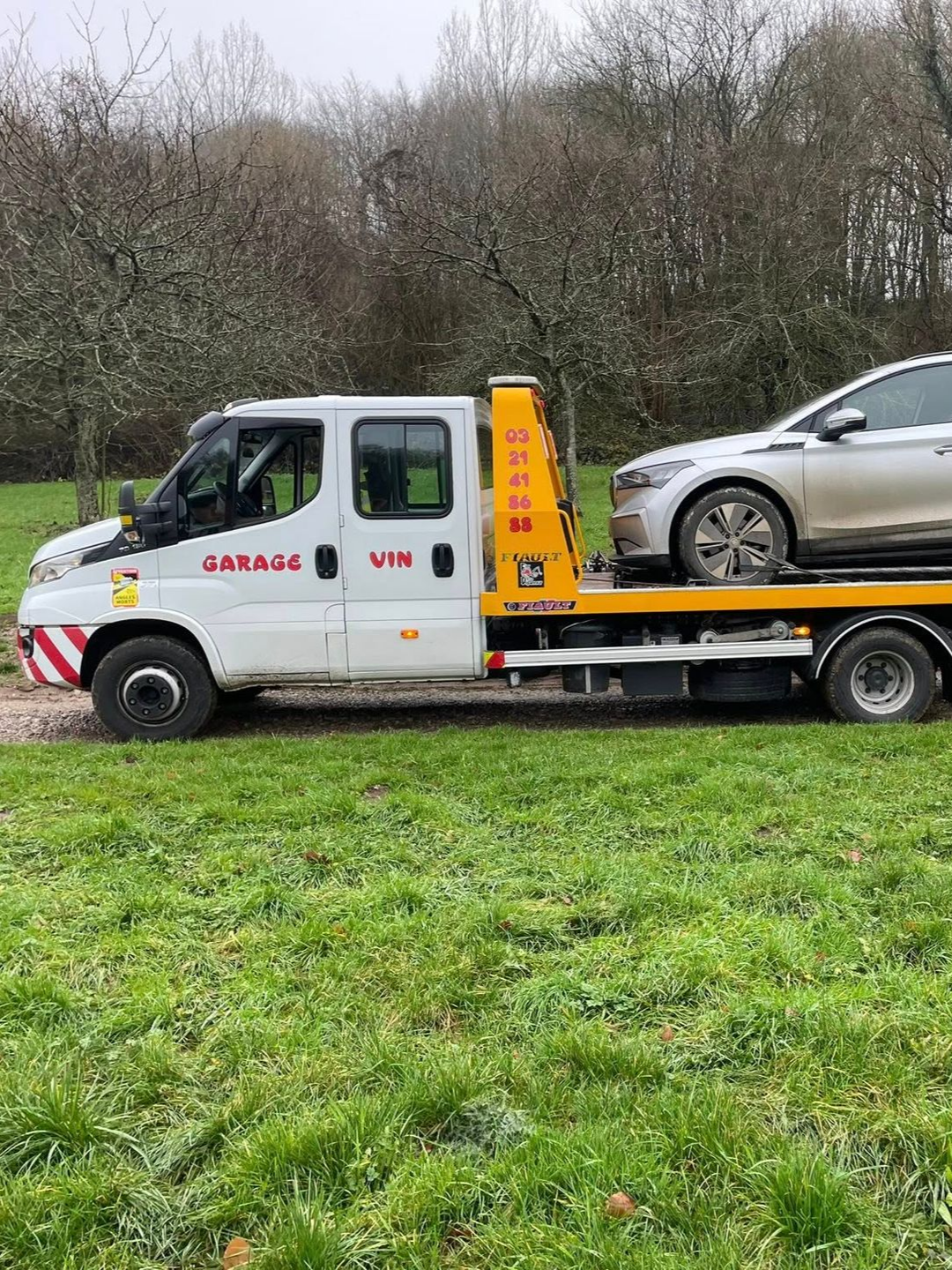 Camion de dépannage blanc avec une voiture chargée sur le plateau, stationné sur l'herbe.