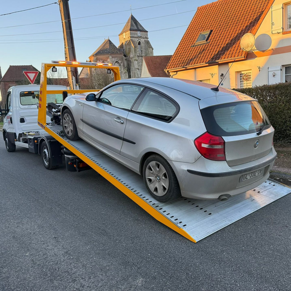 Une voiture argentée chargée sur une dépanneuse à plateau, dans une rue devant des immeubles.