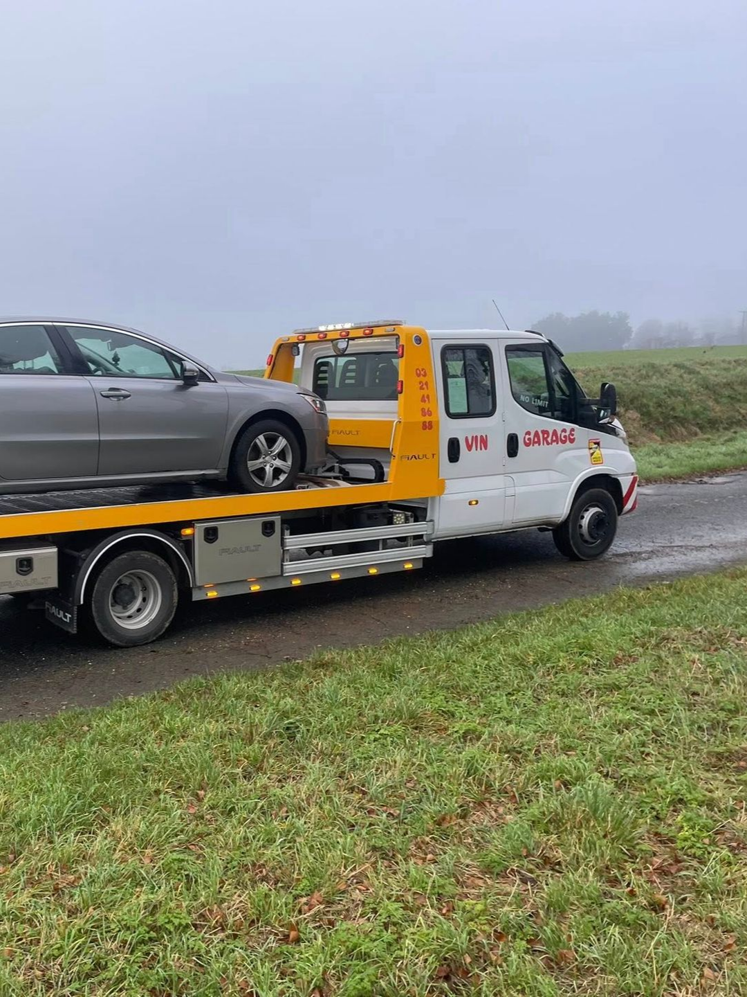 Une dépanneuse transportant une voiture grise sur une route mouillée, entourée de champs, par temps nuageux.
