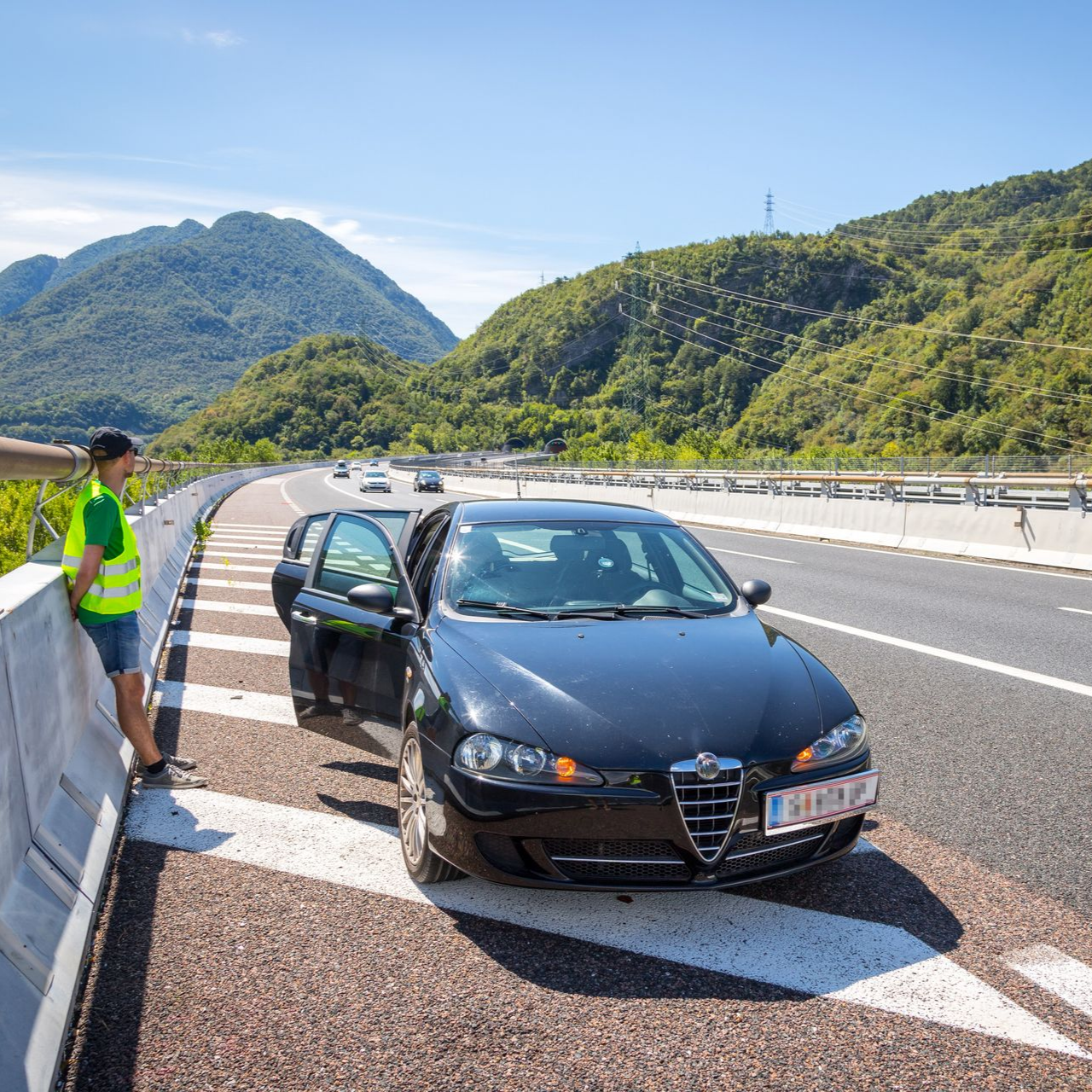 Une personne portant un gilet de sécurité se tient à côté d'une voiture noire dont la portière est ouverte, sur une autoroute avec des montagnes en arrière-plan.