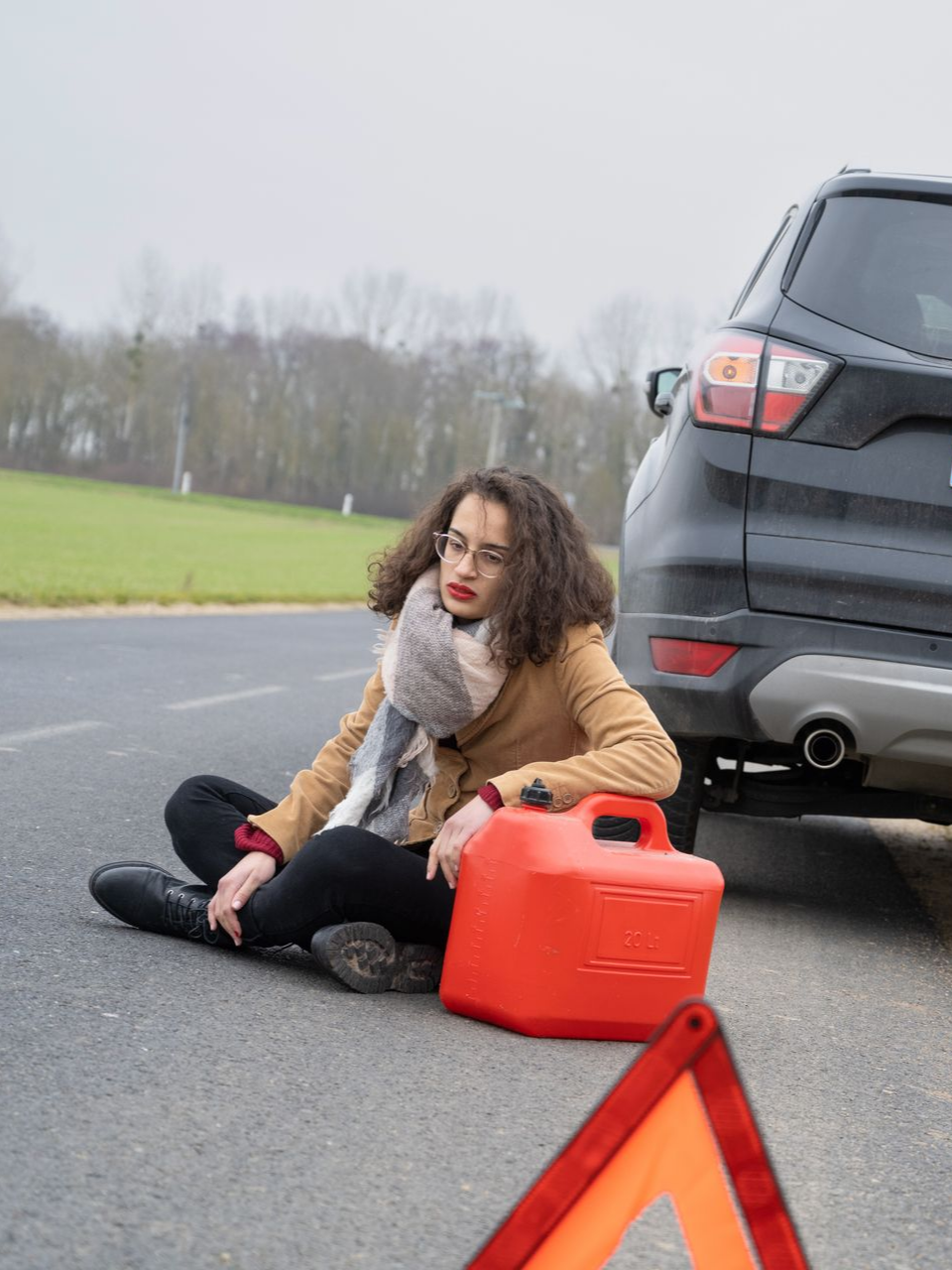 Une femme est assise au bord de la route, à côté d'une voiture, d'un bidon d'essence rouge et d'un triangle de signalisation ; elle semble être en panne.