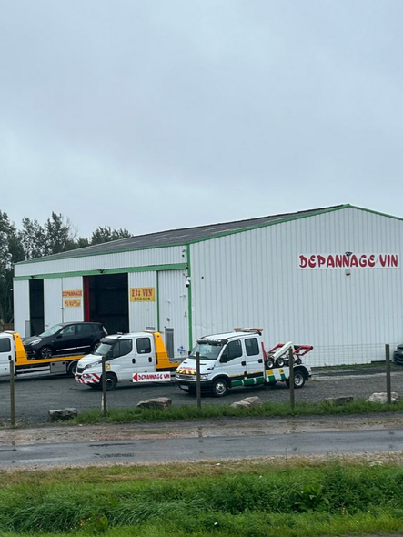 Des dépanneuses sont stationnées devant un bâtiment portant l'inscription DÉPANNAGE VIN. Une voiture noire est en cours de chargement. Ciel couvert.