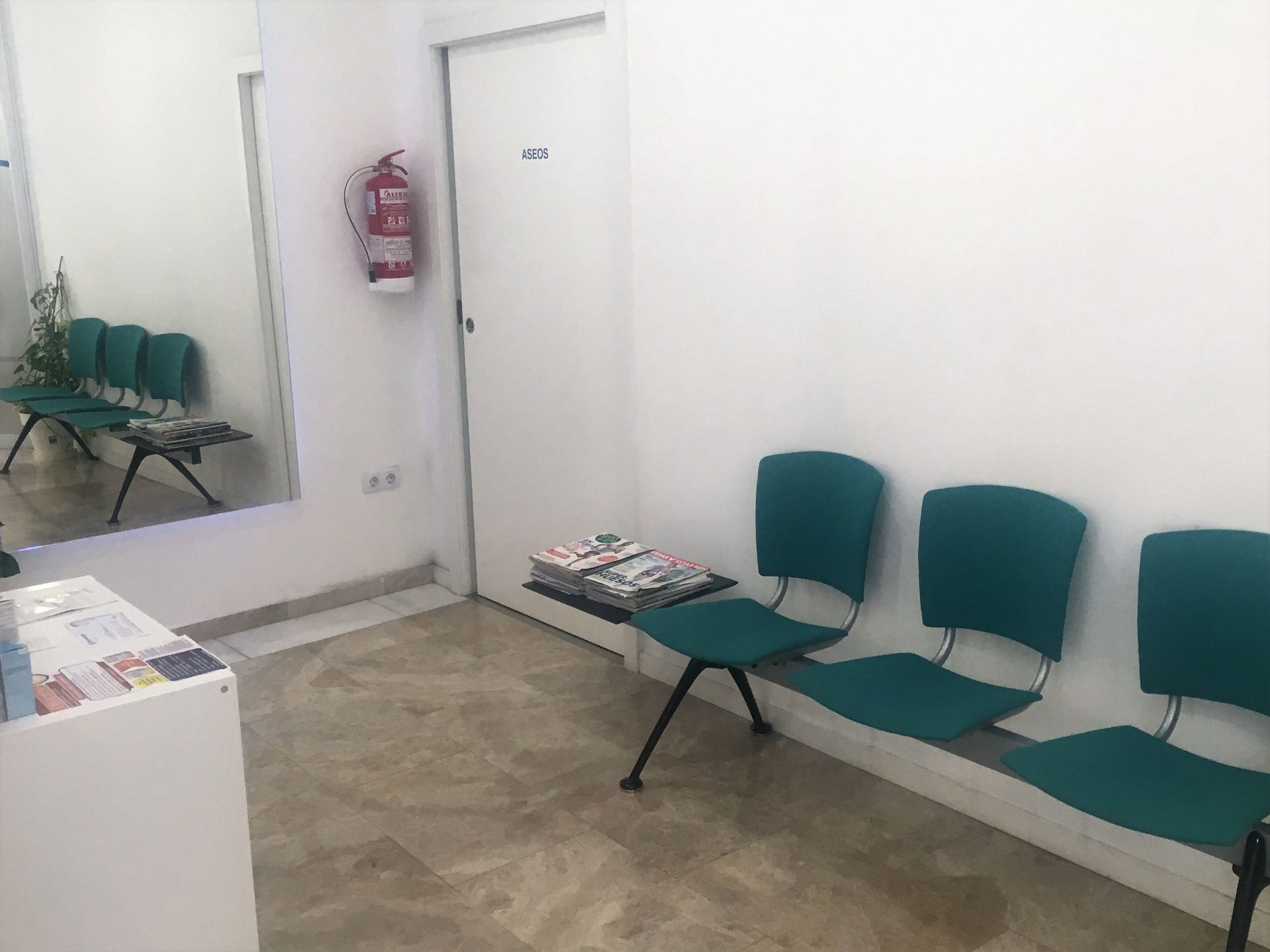 Una sala de espera con sillas verdes y un extintor en la pared.