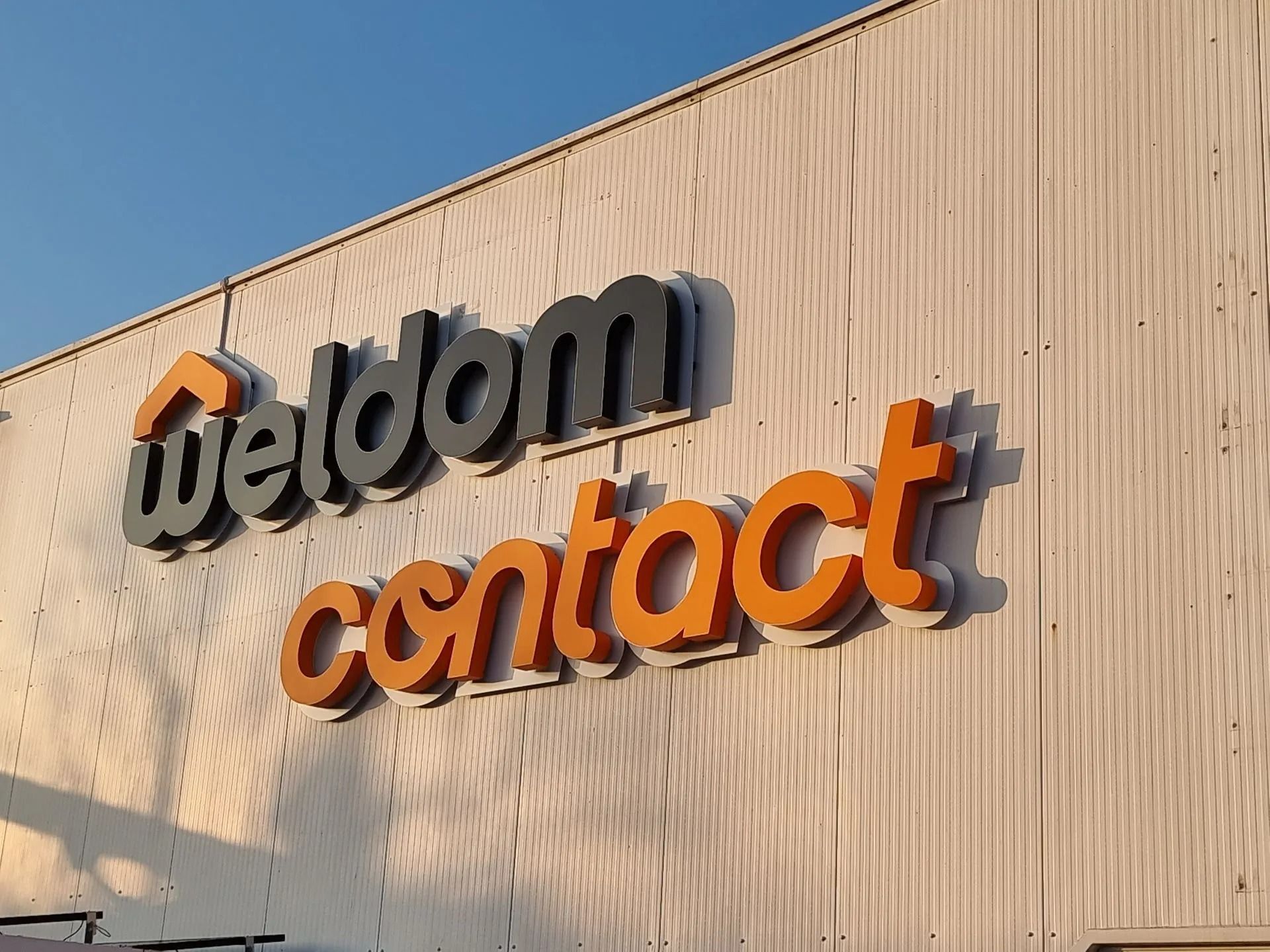 Enseignes Weldom Contact sur une façade en tôle ondulée beige, baignées de soleil.