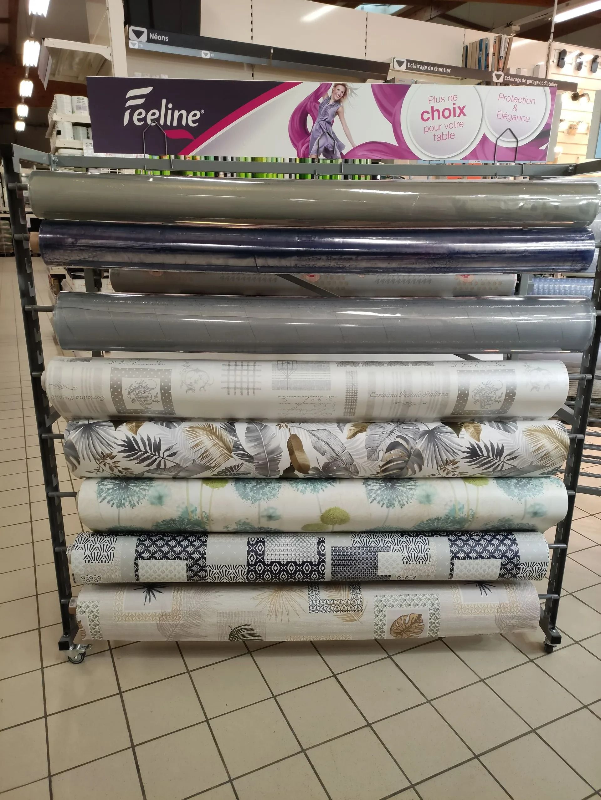 Rouleaux de papier peint décoratif présentés sur un présentoir métallique dans une allée de magasin.