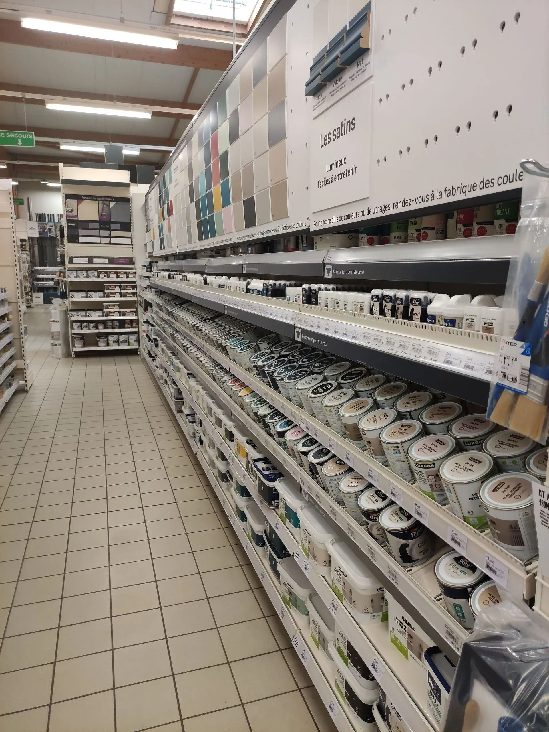 Allée d'un magasin aux longues étagères remplies de pots de peinture et de fournitures, vue au bout du couloir carrelé.