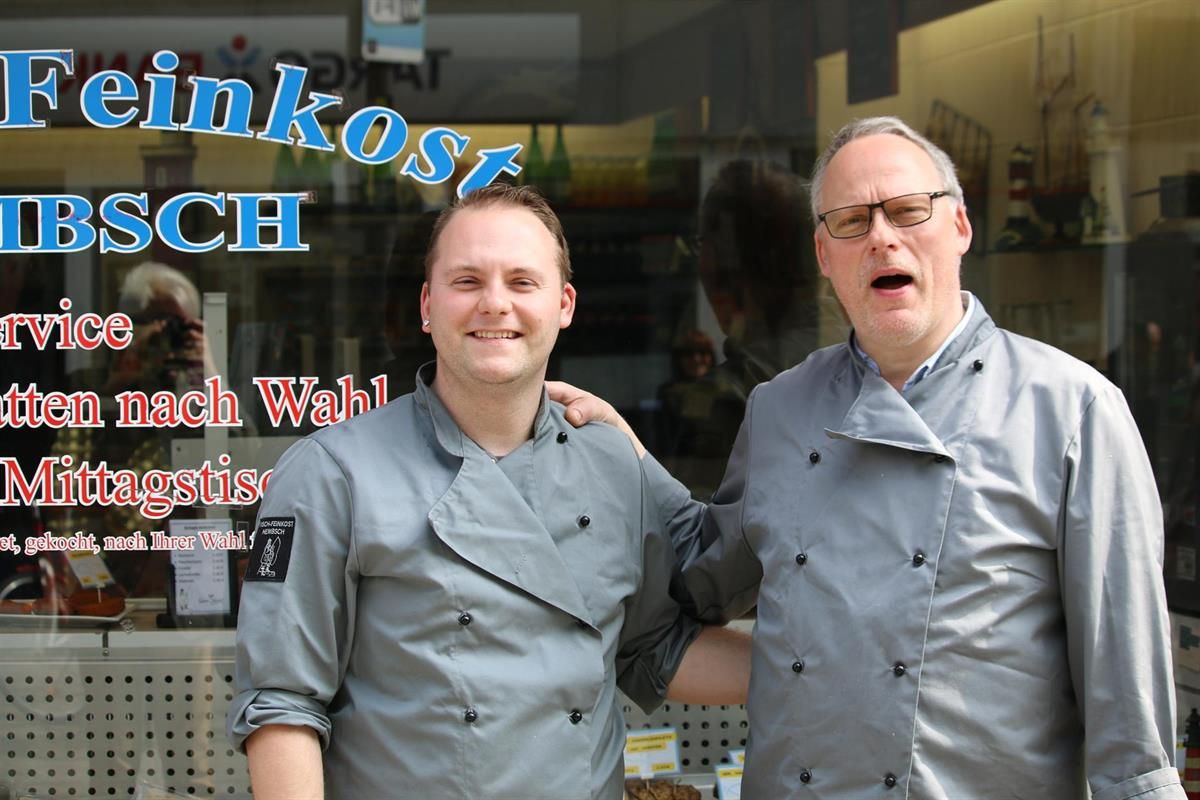 Zwei Männer stehen vor einem Fenster mit der Aufschrift „Feinkost Ibsch“