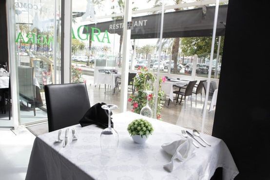 Mesa de restaurante puesta con mantel blanco, servilleta negra, flores y vasos junto a una ventana con vistas a la terraza.