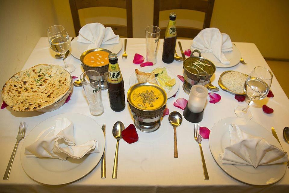 Prepara la mesa del comedor con una variedad de platos indios, vasos, platos y servilletas dobladas bajo una iluminación cálida.