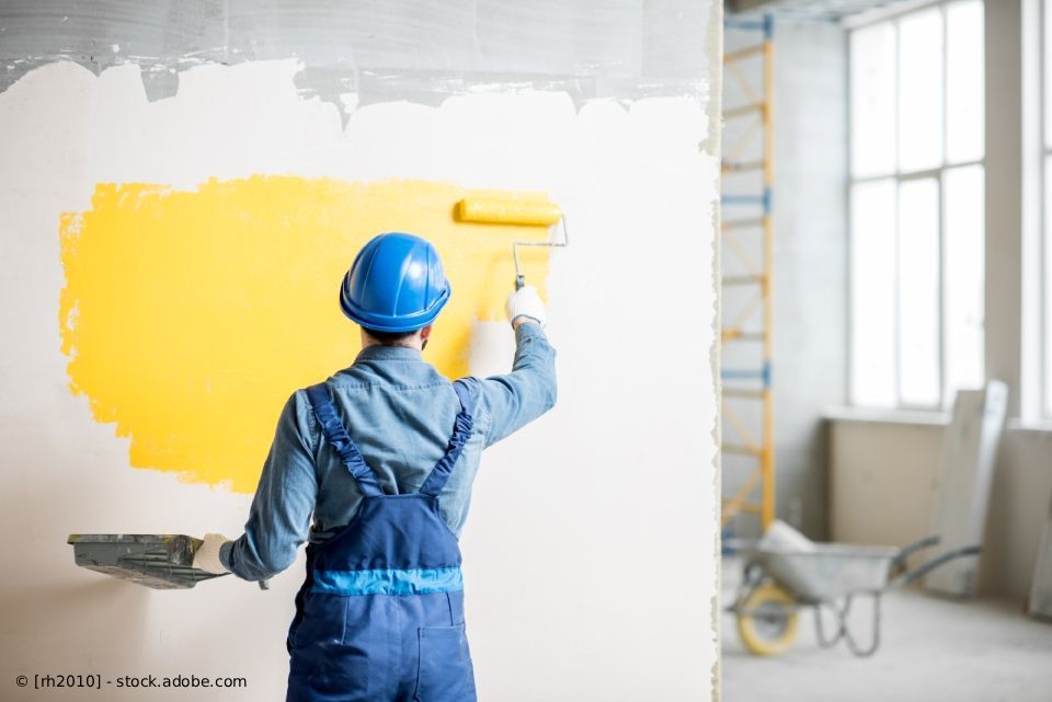 Ein Arbeiter in blauem Overall streicht mit einer Farbrolle eine weiße Wand in Innenräumen gelb.