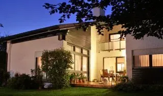 Modernes Haus in der Abenddämmerung, warmes Licht fällt durch große Fenster und davor befindet sich ein Garten.