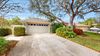 A one-story beige house with a two-car garage, a concrete driveway, and lush landscaping under a sunny sky.