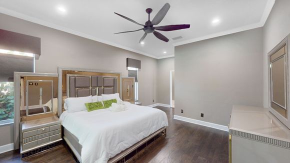 Spacious bedroom with a large bed, dark wood floors, and gray walls. A mirrored dresser and nightstands flank the bed.