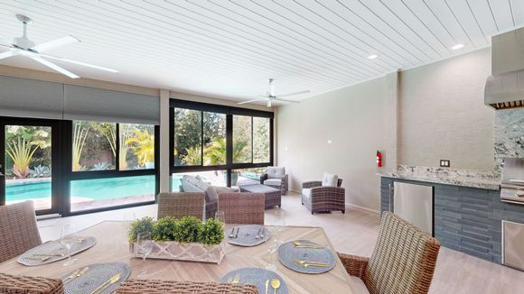 Outdoor dining area with a pool view; a large table is set for a meal. There are rattan chairs and a kitchen with a stone countertop.