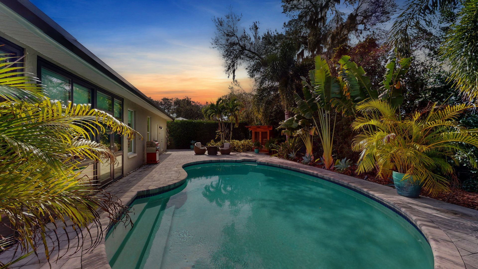 Backyard pool at dusk, surrounded by tropical plants, stone patio, and a glimpse of a house. The sky has orange and blue hues.