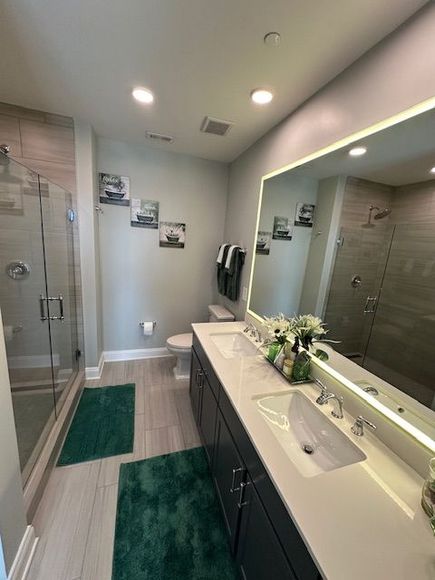 Modern bathroom with a double vanity, large mirror, and glass shower. Green rugs and wall art accent the neutral tones.