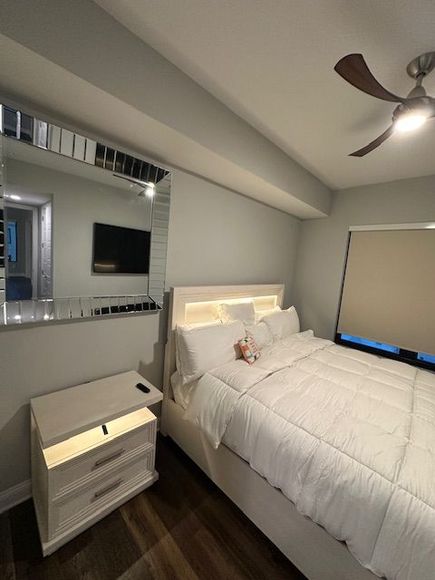 White bedroom with a bed, nightstand, and large mirror. A TV is mounted on the wall and a ceiling fan is visible.