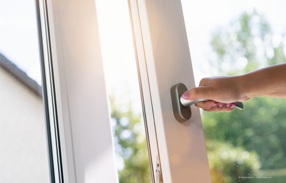 Eine Hand öffnet eine weiße Glasschiebetür, draußen Sonnenlicht und Grün.