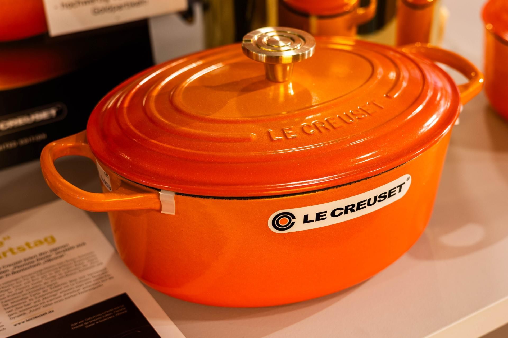 Orangefarbener Le Creuset Dutch Oven mit Deckel und Seitengriffen. Goldener Knopf, weißes Etikett.