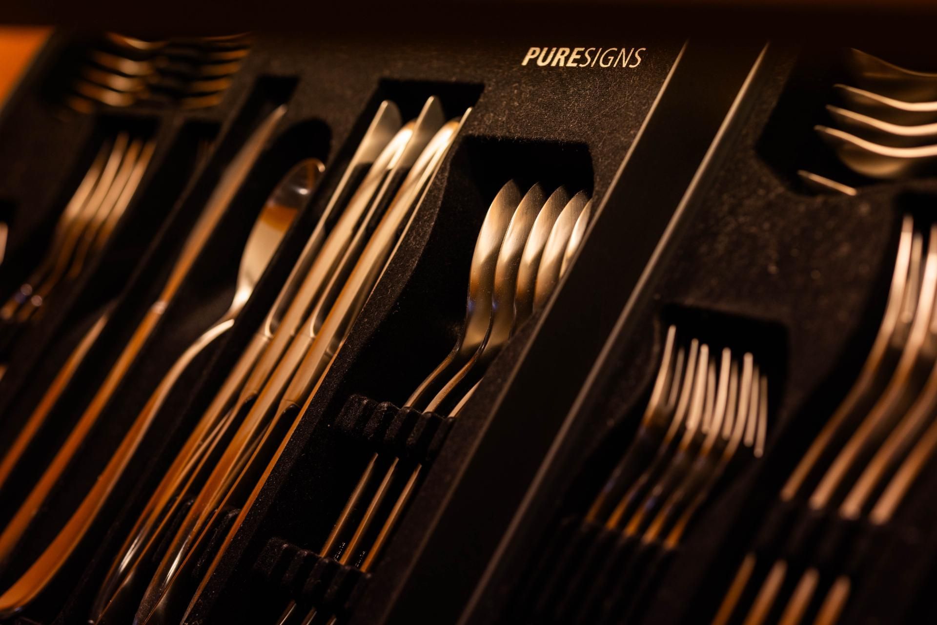 Edelstahl-Besteckset, ordentlich angeordnet in einer schwarzen Aufbewahrungsschale. Das Etikett „PURETENS“ ist sichtbar.