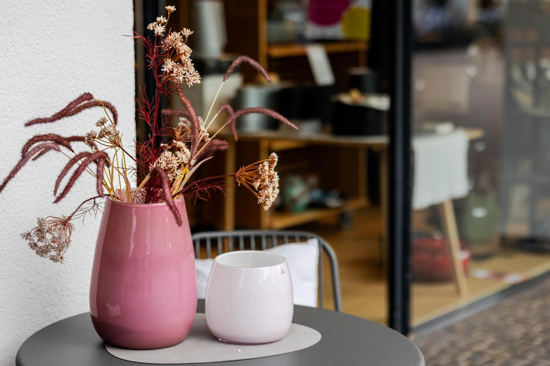 Zwei rosa Vasen mit getrockneten Blumen auf einem grauen Tisch im Freien, im Hintergrund ein Schaufenster.