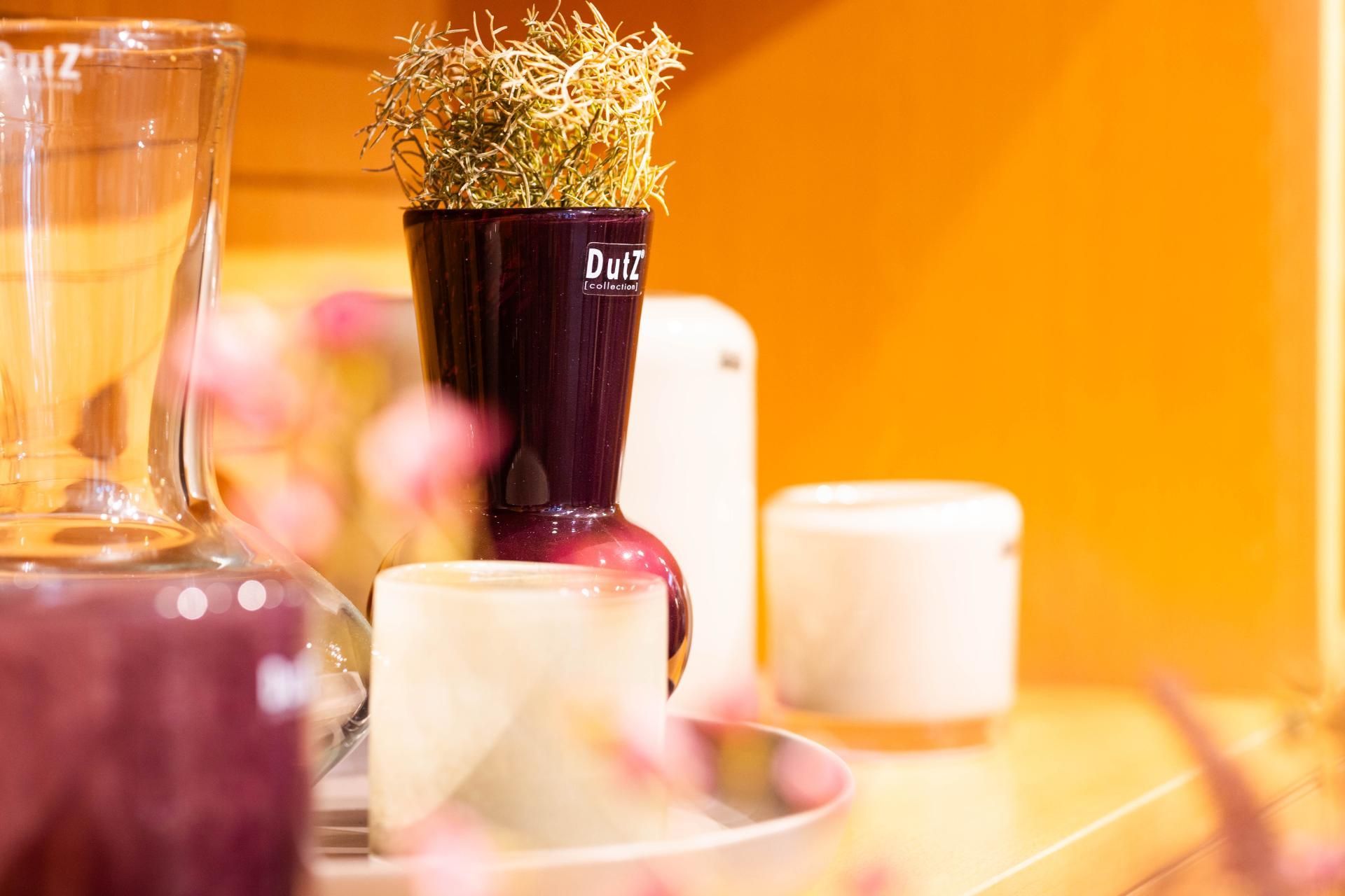 Stillleben mit Dekorationsgegenständen auf einem Regal: lila Vase mit Grünpflanzen, Kerzen und eine Karaffe.