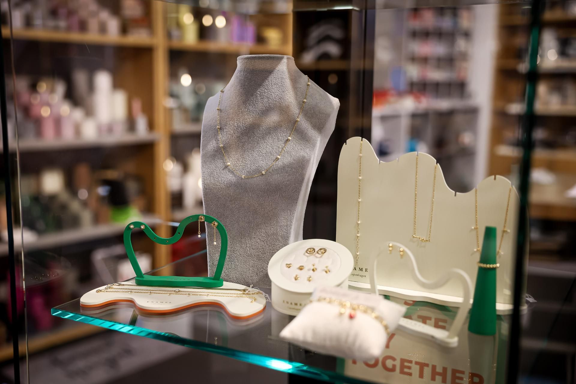 Schmuckvitrine mit verschiedenen Halsketten, Ohrringen und Ringen auf weißen und grünen Ständern; Regale im Hintergrund.