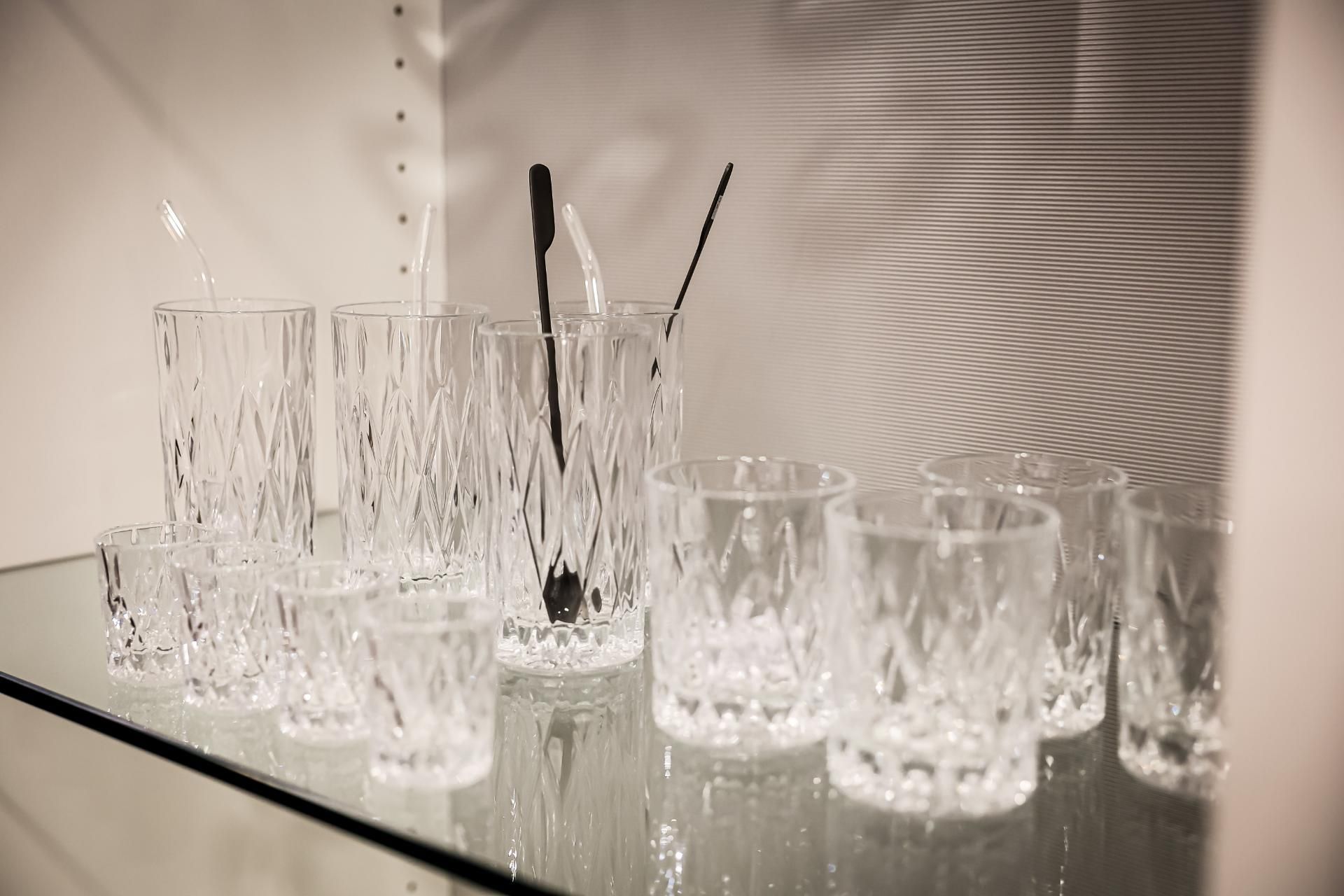 Auf einem Regal ausgestellte Glaswaren: mehrere Kristallgläser in diversen Größen, Strohhalme und Longdrink-Rührlöffel.