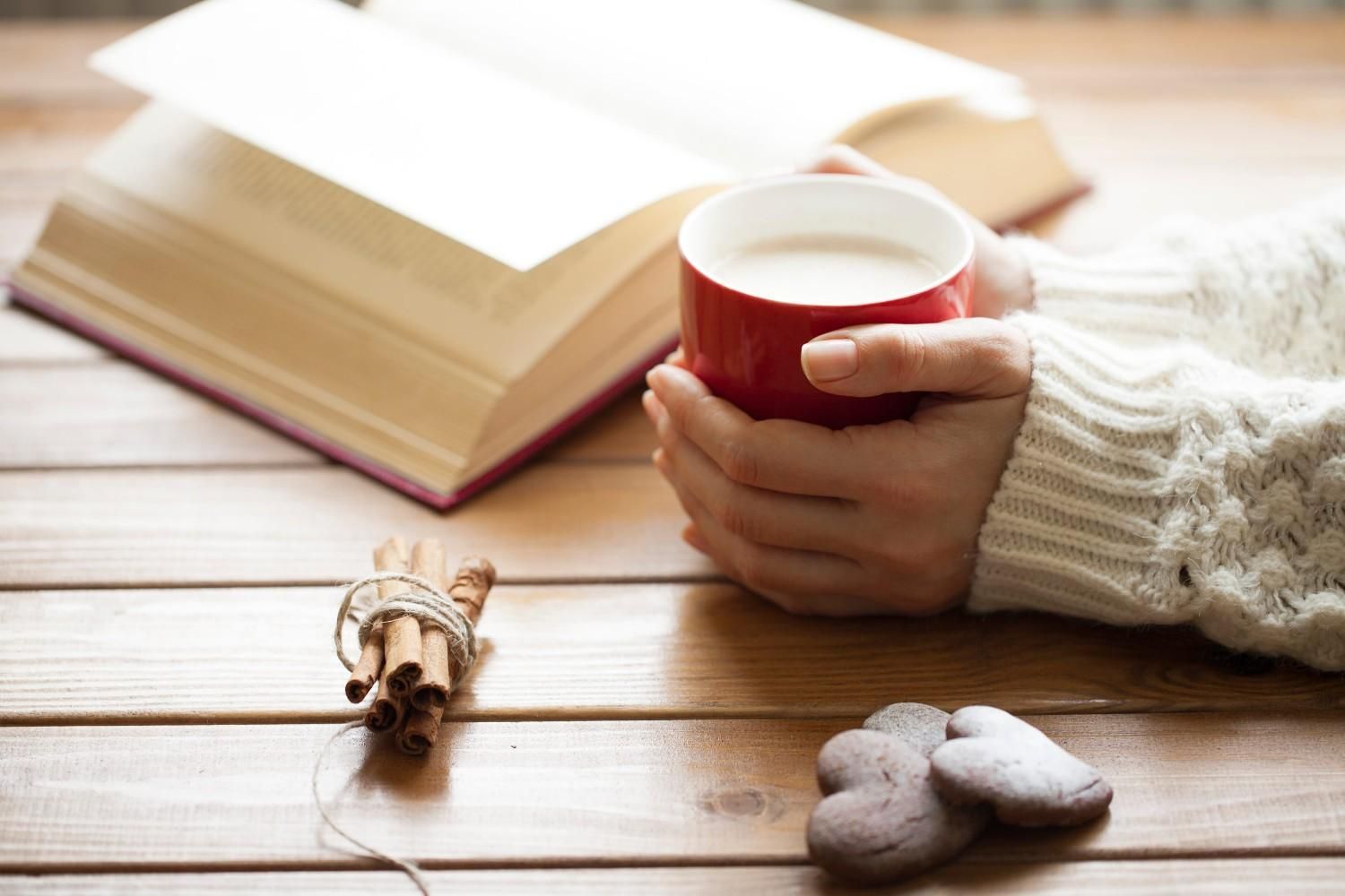 Una mano sostiene una taza roja junto a un libro abierto sobre una mesa de madera, con un pequeño corazón decorativo cerca.