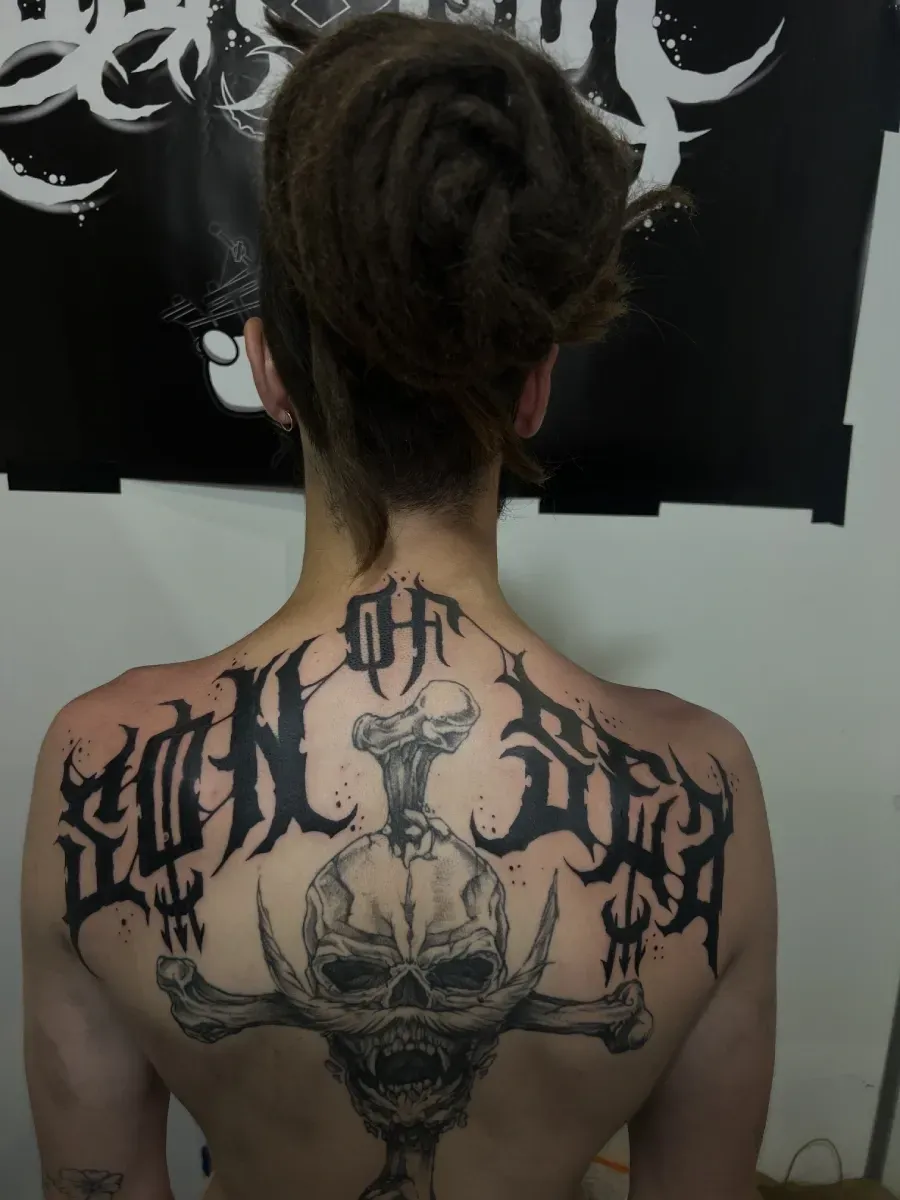 Tatuaje en la espalda: Calavera y huesos cruzados con letras góticas, persona con rastas.