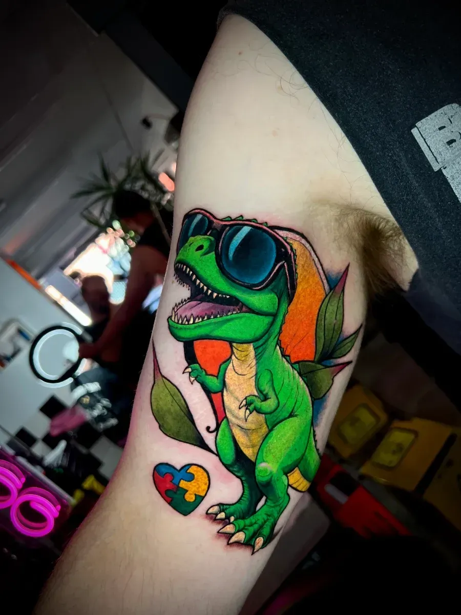 Tatuaje de dinosaurio verde con gafas de sol, corazón de rompecabezas autista y hojas en el brazo.