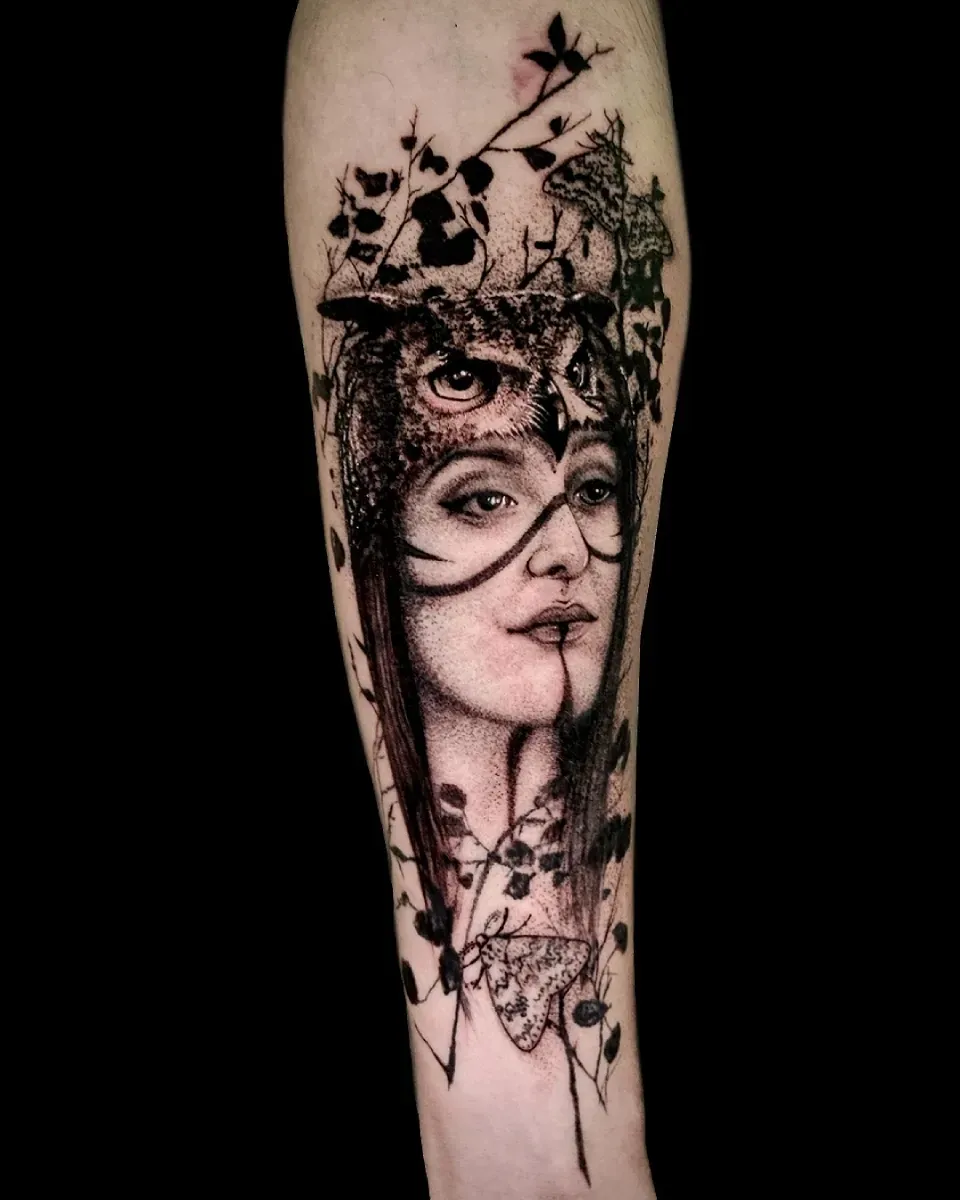 Tatuaje en el brazo del rostro de una mujer con una máscara de búho, rodeada de follaje. Blanco y negro.