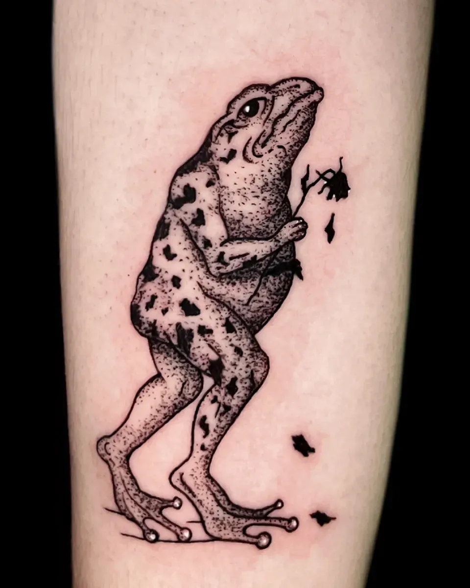 Tatuaje de una rana moteada, de pie y sosteniendo una flor marchita.
