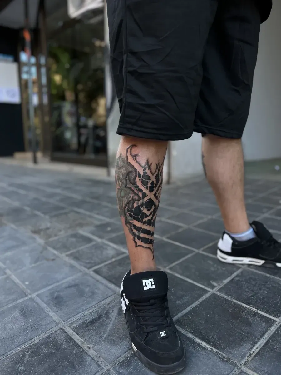 Pierna de un hombre con un tatuaje, vestido con pantalones cortos negros y zapatos DC, de pie sobre una acera de baldosas.