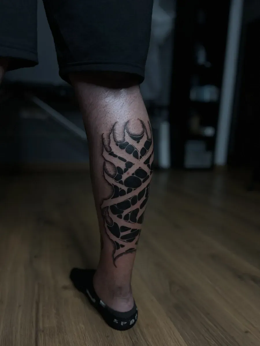 Tatuaje tribal negro en la pantorrilla. La persona está de pie sobre un suelo de madera, con pantalones cortos y zapatos negros.