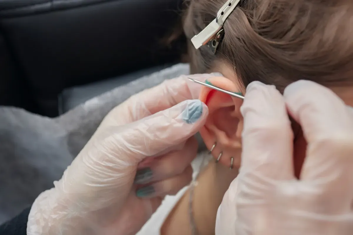 Persona que se perfora la oreja, con las manos enguantadas sosteniendo la aguja perforadora en la oreja.