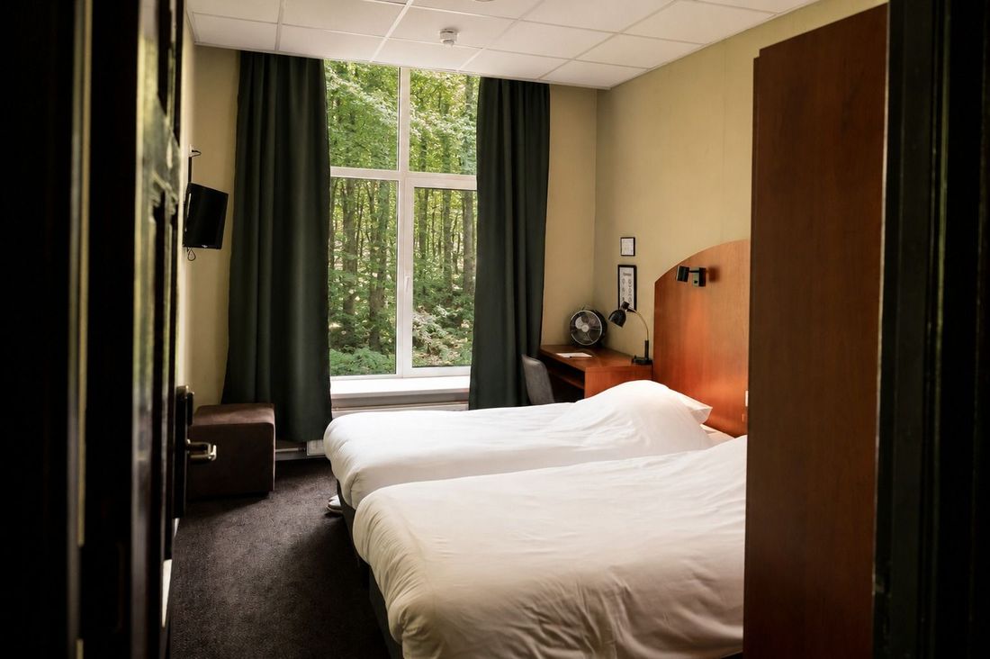 Tweepersoonskamer met een bed bij het raam, een donkerhouten deur en groene gordijnen met uitzicht op bomen.