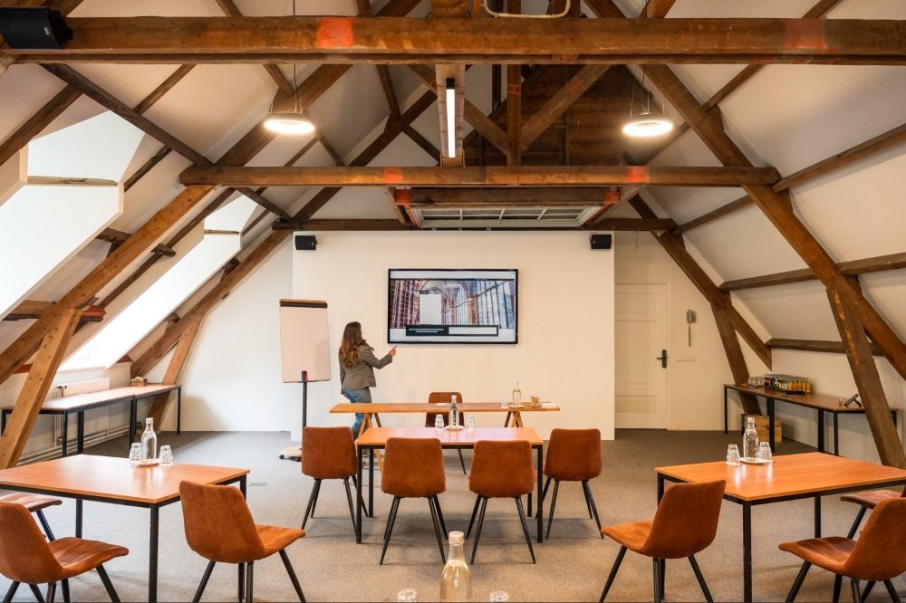 Moderne vergaderruimte met een persoon achter een whiteboard, rijen tafels en zichtbare houten balken.