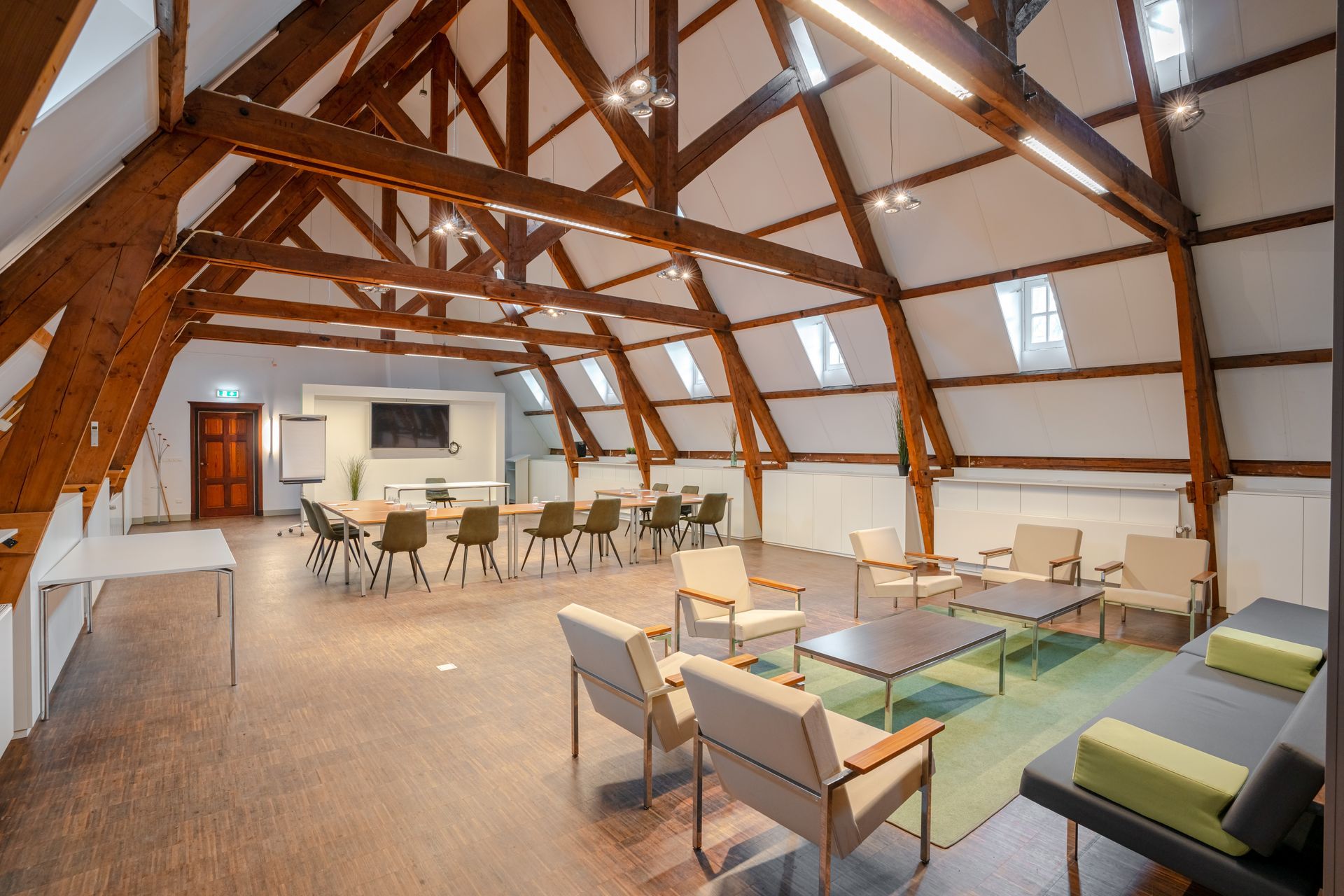 Ruime vergaderzaal met houten balken, banken, fauteuils en een eethoek onder sfeervolle verlichting.