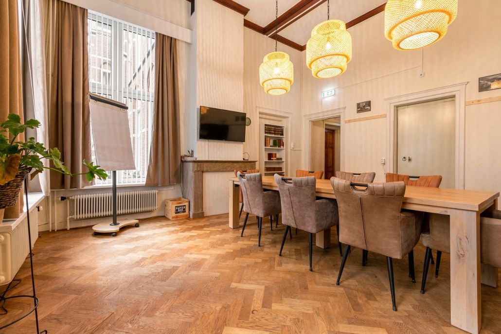 Lichte vergaderruimte met houten tafel, stoelen, grote ramen en hangende, geweven pendellampen.