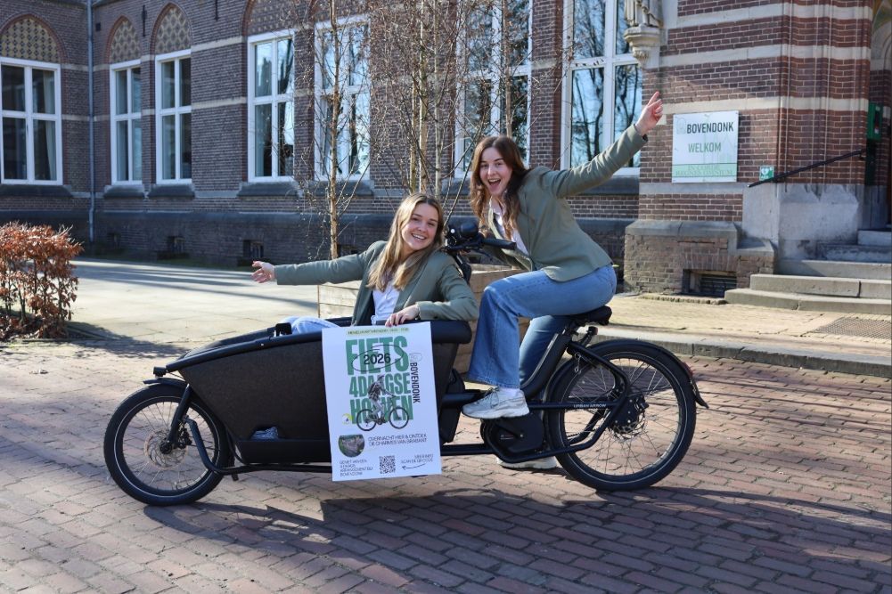 Fietsers rijden op een met bomen omzoomd pad tijdens Fiets4Daagse Hoeven, met evenementlogo en tekstoverlay.