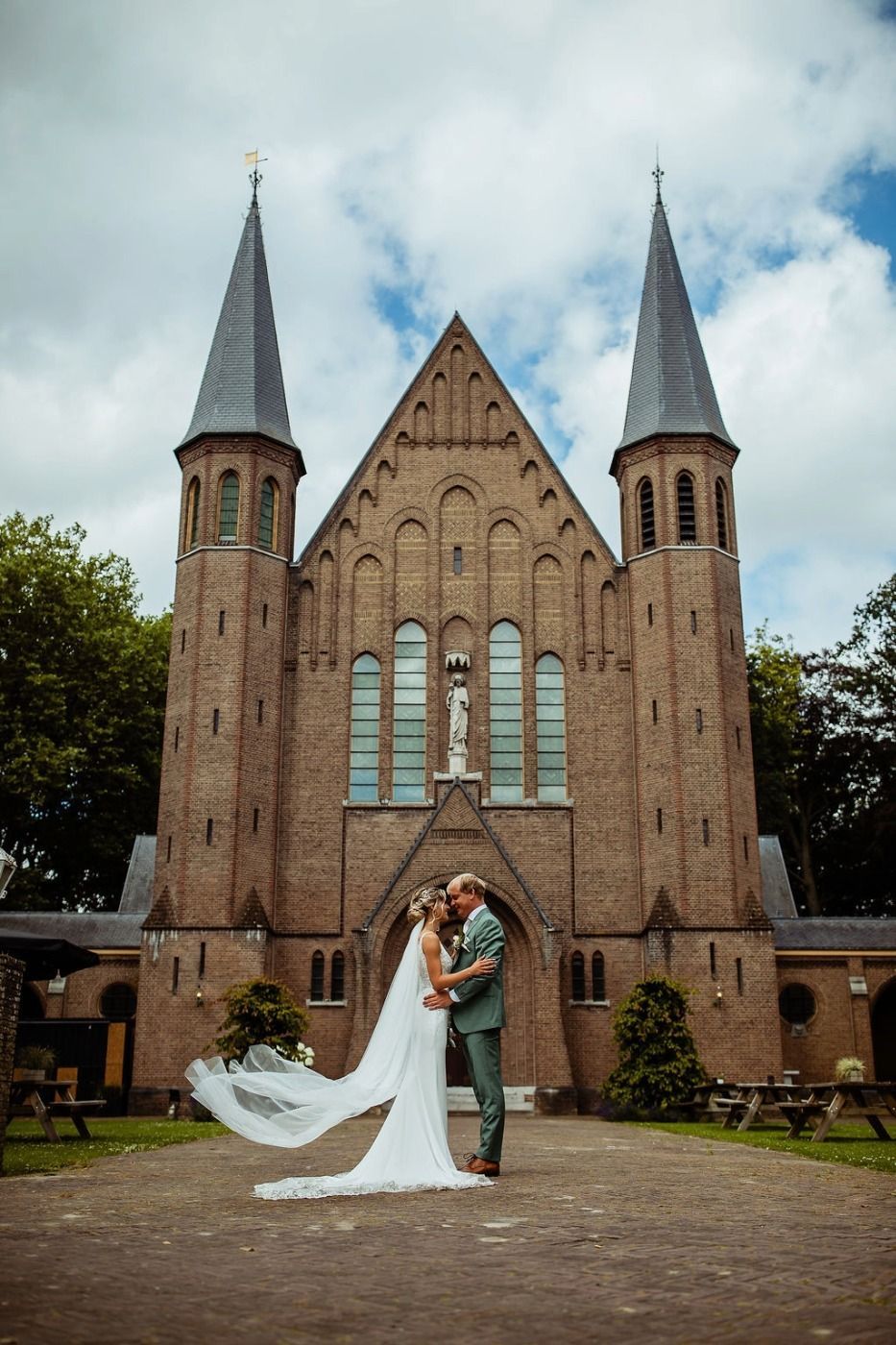 Bruid en bruidegom omhelzen elkaar voor de Kapel, terwijl de sluier van de bruid in de wind wappert.