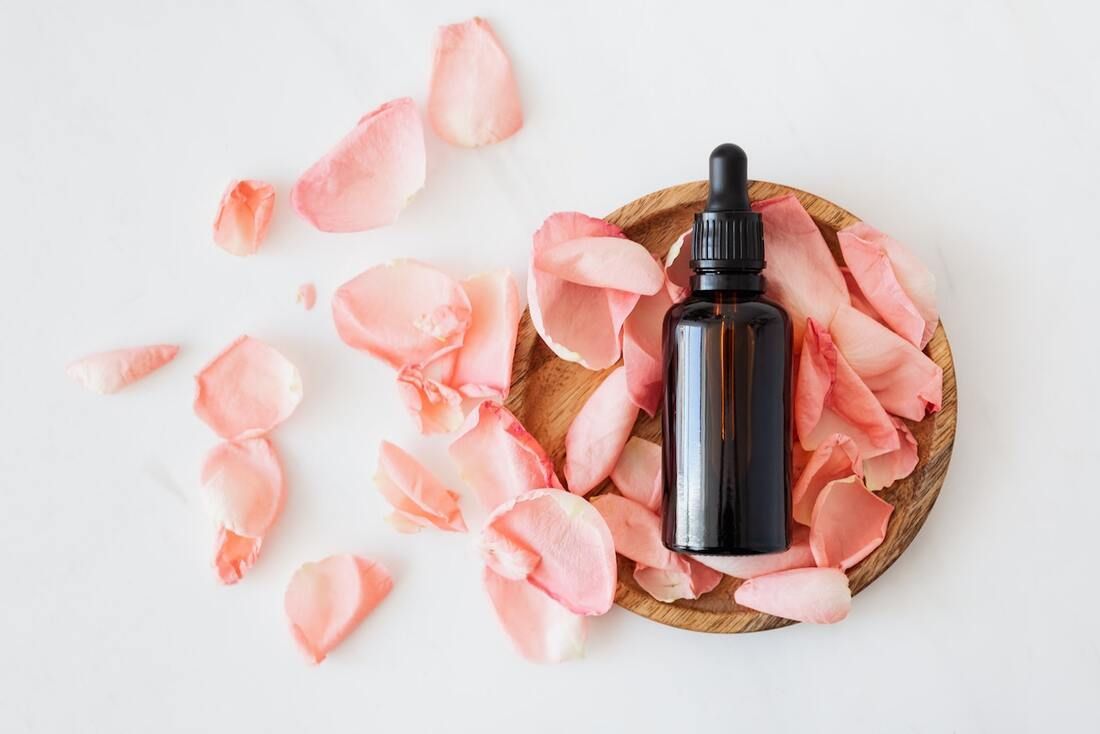 Una botella de aceite esencial se encuentra en un recipiente de madera rodeado de pétalos de rosa rosa.