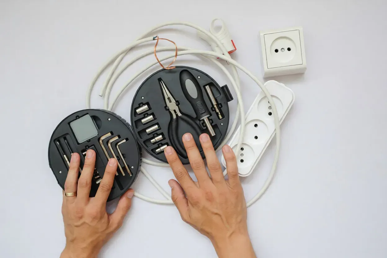 Manos trabajando en dos juegos de herramientas circulares, cable eléctrico, un enchufe de pared y una regleta sobre un fondo blanco.