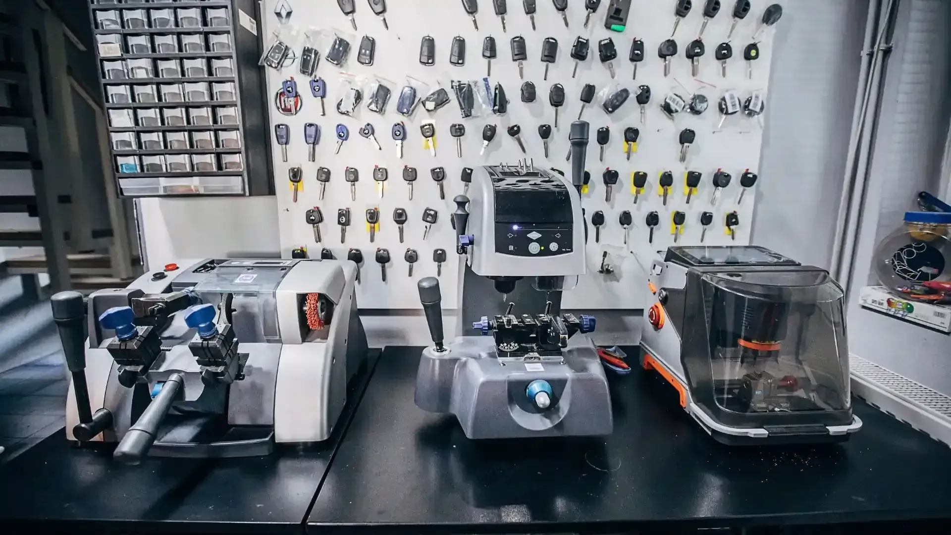 Tres máquinas duplicadoras de llaves se encuentran sobre un banco de trabajo, con un panel montado en la pared que muestra numerosas llaves al fondo.