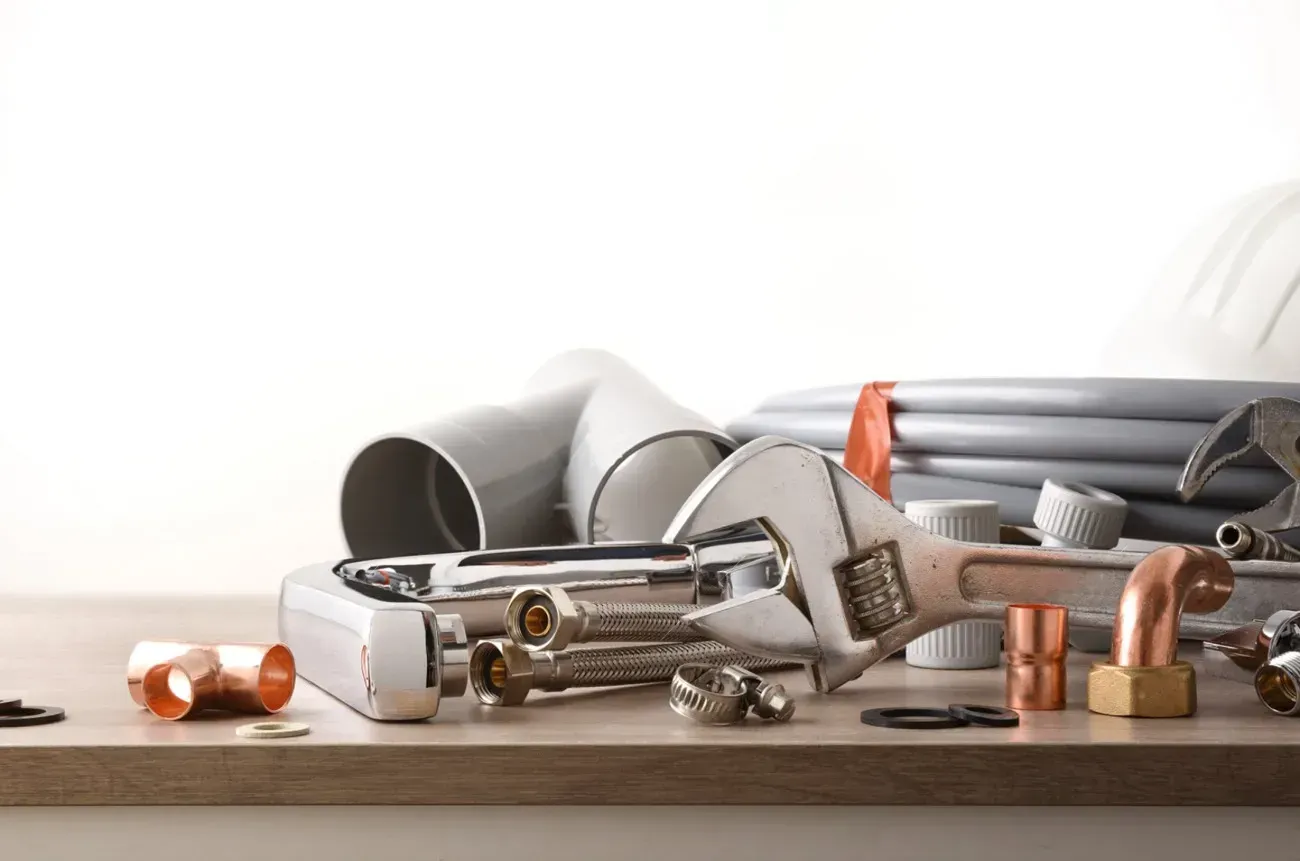 Herramientas de fontanería y diversos accesorios de tubería de metal y plástico dispuestos sobre una superficie de madera sobre un fondo blanco.